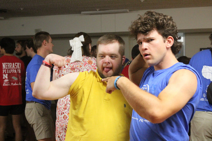 Two young men flexing biceps in a casual indoor setting with other people in the background.